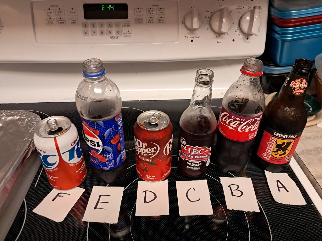 An assortment of cherry-flavored sodas arranged on a kitchen counter, labeled from A to F with paper tags, including Cherry Dr. Pepper, Cherry RC, Cherry Coke, Wild Cherry Pepsi, IBC Cherry Soda, and Sprecher Cherry Cola.